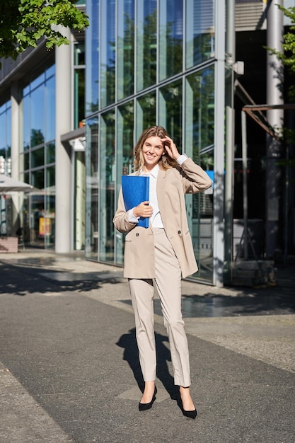 Free photo vertical shot of businesswoman with folder standing on a street near office buildings wearing suit f