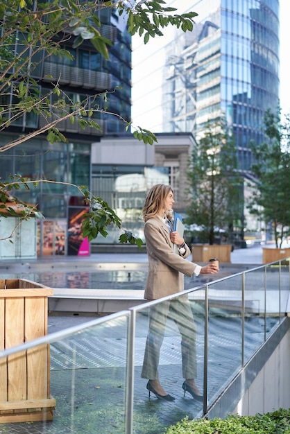 Free photo vertical shot of corporate woman in beige suit standing outside on street drinks morning coffee take