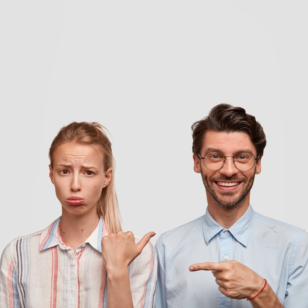 Free Photo vertical shot of dissatisfied blonde woman purses lower lip, being offended by guy. positive unshaven hipster with pleasant smile, points at girlfriend, isolated over white wall, free space