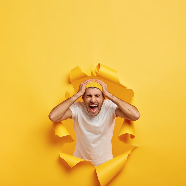 Free Photo vertical shot of emotional guy touches head with both hands, wears casual white t shirt, stylish yellow hat