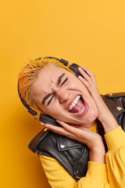 Free Photo vertical shot of female hipster meloman with dyed hair tilts head sings song along listens music in wireless headphones wears fashionable outfit enjoys rock playlist isolated over yellow wall