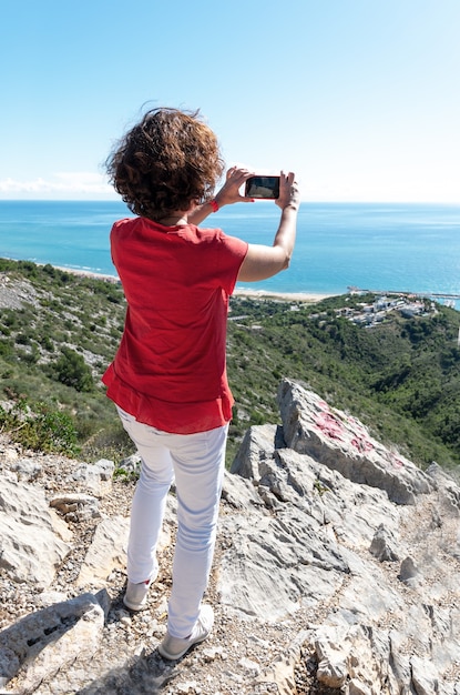 Vertical shot of a female standing on rocks and photographing the beautiful sea