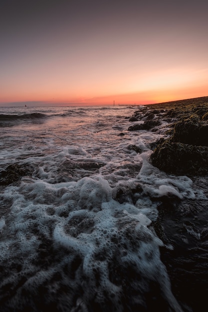 Vertical shot of foamy waves of the sea coming to the shore with the amazing sunset