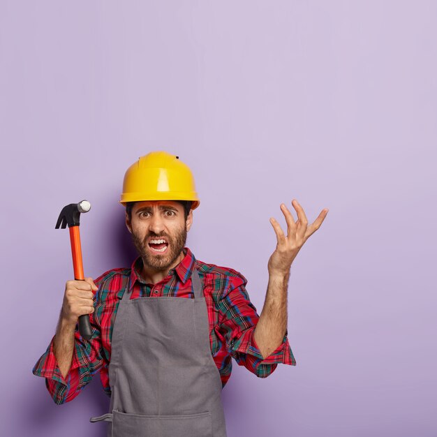 Free photo vertical shot of frustrated repairman has puzzled face expression, hammer in hand, raises arm with indignation, dressed in workwear, cannot understand what to repair