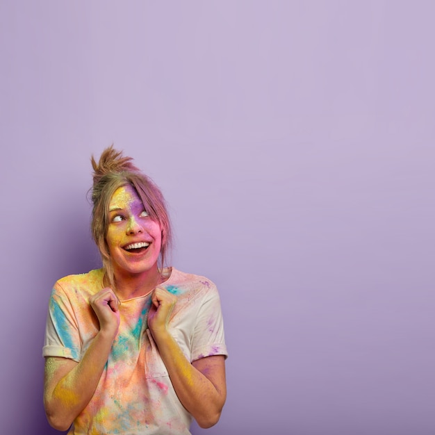 Vertical shot of glad female with bright colorful mix of paints, raises clenched fists, enjoys traditional hindu festival celebration in India, wears casual white t shirt, focused above on free space