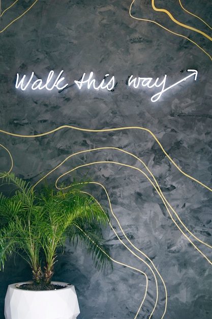 Vertical shot of a green plant in a white pot near a gray wall with a neon sign