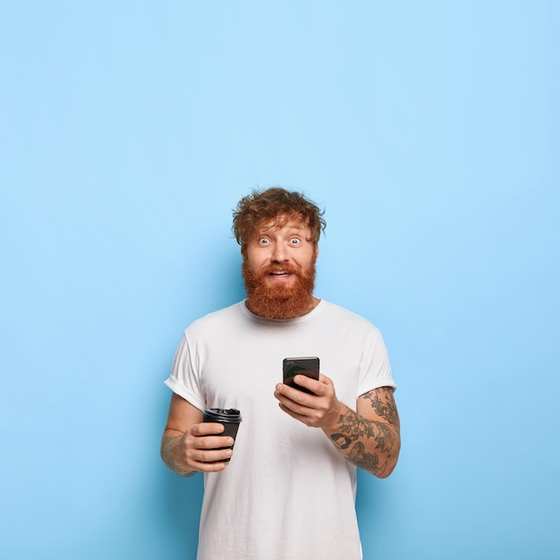 Vertical shot of handsome cheerful red haired guy posing with his phone