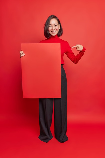 Vertical shot of happy Asian woman with dark hair pleasant smile points at square paper for template shows mockup for your design advertises promo banner dressed in stylish clothes poses indoor