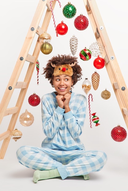 Free photo vertical shot of happy curly haired woman dressed in checkered pajama sleepmask sits crossed legs on floor decorates house with new year toys dreams about something believes in miracle on christmas