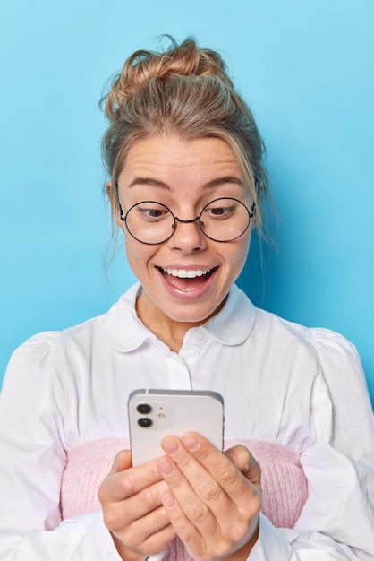 Free photo vertical shot of happy surprised excited woman looks at smartphone screen receives pleasant notification watches video satisfied with new application smiles from amazement isolated on blue wall