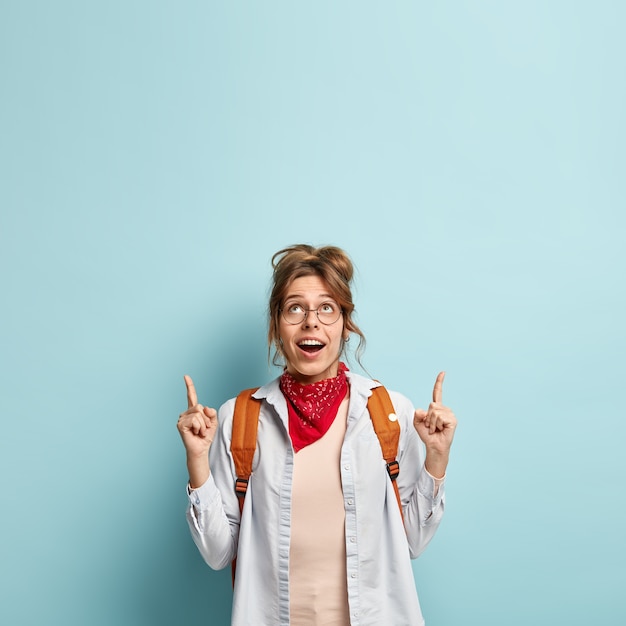 Free photo vertical shot of happy young female points above with both fore fingers, shows awesome product on top, wears stylish casual clothes