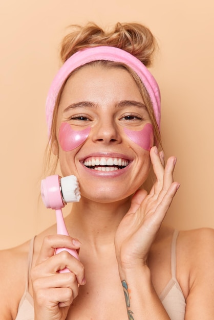 Free photo vertical shot of happy young woman touches face gently smiles broadly shows white teeth uses massager applies hydrogel patches under eyes to remove fine lines isolated over beige background.