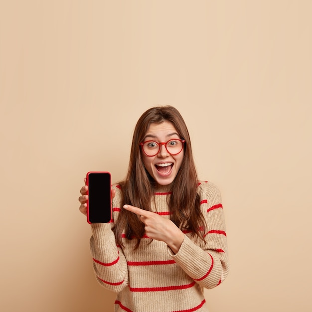 Free photo vertical shot of joyful caucasian female points at smart phone device, shows black blank screen for your advertising text, laughs, has pleased expression, isolated over beige wall with free space