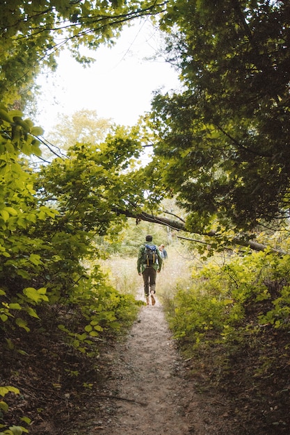 Free Photo vertical shot of a male with a backpack walking on a narrow pathway in middle of trees and plants