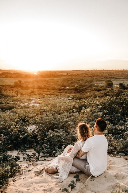 Vertical shot of a man and a woman sitting and hugging in a field while looking at the sunset