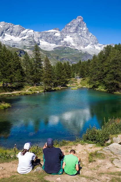 Vertical shot of a people sitting near a pond surrounded by trees with a mountain in the distance