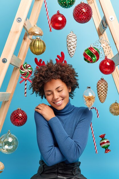 Vertical shot of pleased African American woman enjoys festive magical atmosphere at home wears comfortable turtleneck poses 