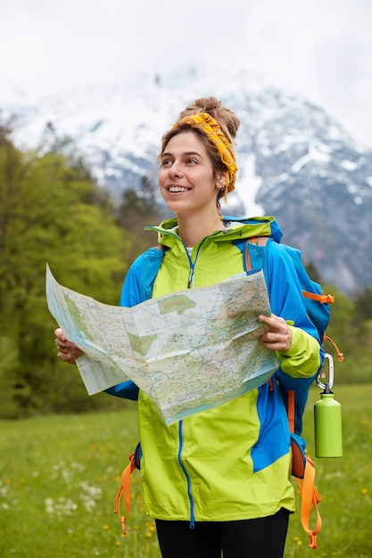 Vertical shot of pleased Caucasian female traveler carries travel map, dressed in bright anorak