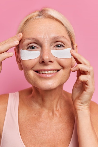 Vertical shot of pleased forty years old blonde woman applies undereye patches provides intensive hydration smiles gently reduces signs of aging has healthy skin poses against pink background.