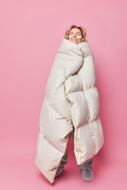 Vertical shot of pleased young woman feels warm as wrapped in soft blanket stands with closed eyes applies beauty patches poses in full length against pink background. Morning time and awakening