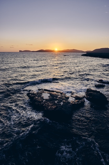 Vertical shot of rocks in the sea with the sun shining behind the mountains in the distance