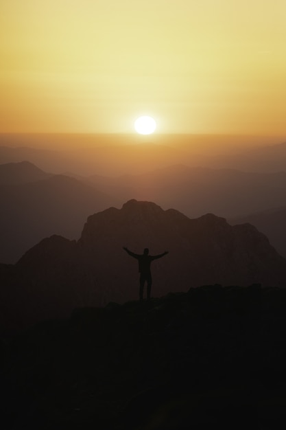 Vertical shot of the silhouette of a male tourist in top of the mountain looking at the sunset