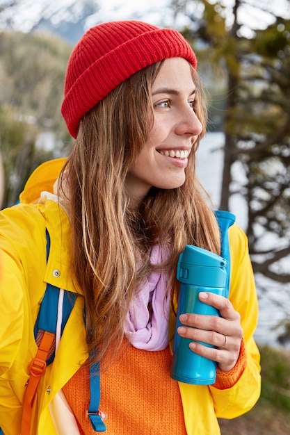 Free photo vertical shot of smiling female traveler wears red hat, yellow coat, stretches hand, makes selfie with unrecognizable device