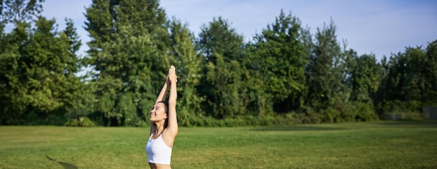 Free photo vertical shot of smiling korean woman doing tree yoga asana stretching on rubber mat in park