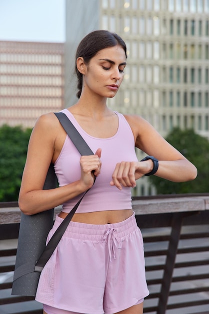 Vertical shot of sportive slim young woman in activewear checks time on smartwatch waits for trainer to start fitness training or yoga practice poses at bridge