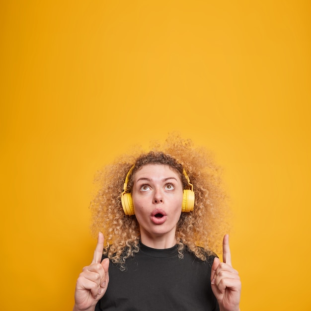 Free Photo vertical shot of surprised wondered young woman with curly hair points above shows amazing advertisement listens music via headphones wears casual yellow t shirt isolated over yellow wall