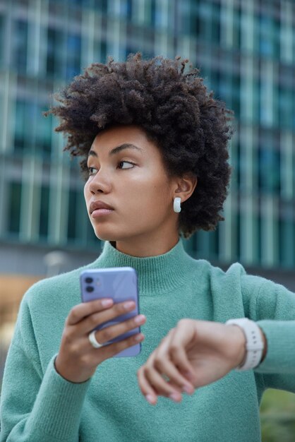 Vertical shot of thoughtful curly haired young woman waits for someone checks time on watch holds mobile phone sends sms to friend wears casual jumper poses outside against blurred background