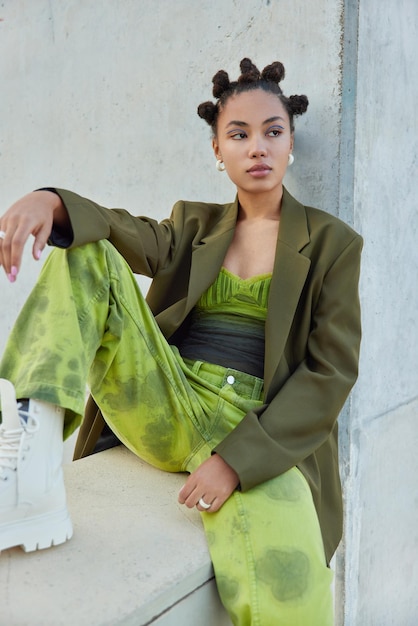 Free Photo vertical shot of thoughtful fashionable woman dressed in green clothes and white boots sits near grey concrete wall has bun hairstyle vivid makeup thinks about something urban lifestyle concept