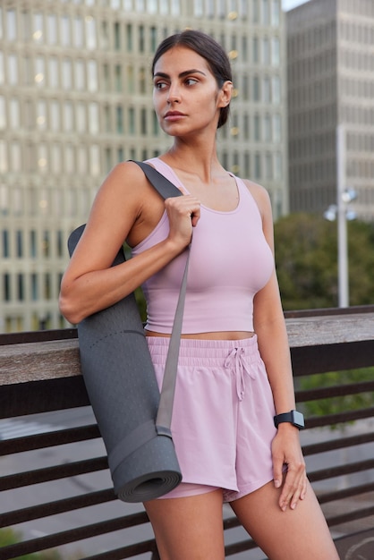 Free photo vertical shot of thoughtful sporty woman dressed in tracksuit carries fitness mat concentrated away into distance poses outdoors against blurred urban background. athletic female going to have pilates