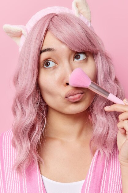 Vertical shot of thoughtful young Asian woman uses brush applies makeup powder on nose keeps lips folded concentrated away has long dyed hair wears headband poses indoor. Cosmetic procedures