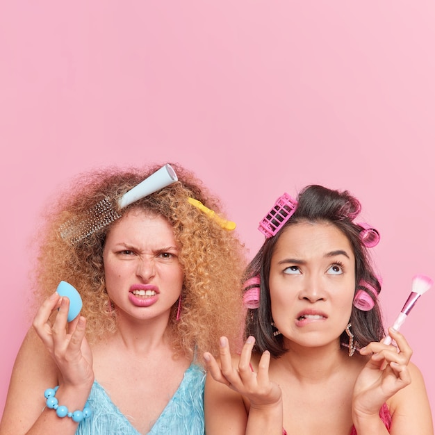 Free Photo vertical shot of two displeased women being in hurry get prepared for party do make up use cosmetic tools smirk face apply rollers comb hair pose together against pink wall with copy space above