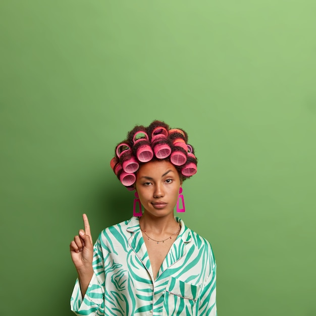 Free photo vertical shot of unimpressed dark skinned woman wears hair curlers and indicates on copy space, shows blank space against green wall for your promo, cares about appearance, prepares for date