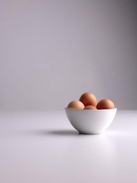 Free photo vertical shot of a white bowl with brown eggs in it on a white surface and grey clean background