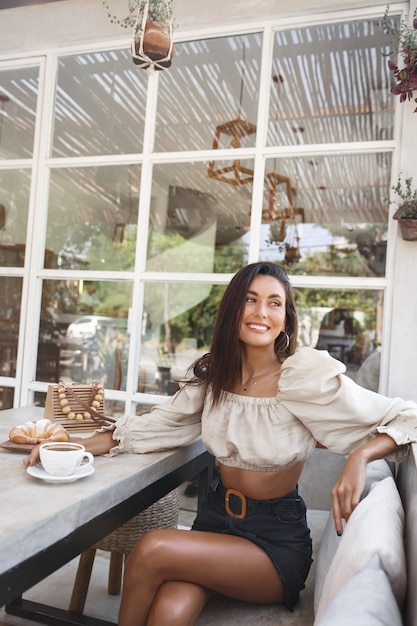 Free Photo vertical shot of a woman in fashionable outfit sitting cafe area with coffee and croissant, looking away at the street.