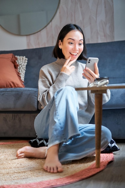 Vertical shot of young asian girl looks surprised at her mobile phone reads amazing news notificatio