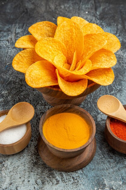 Vertical tasty potato chips decorated like flower shaped different spices with spoons on gray table