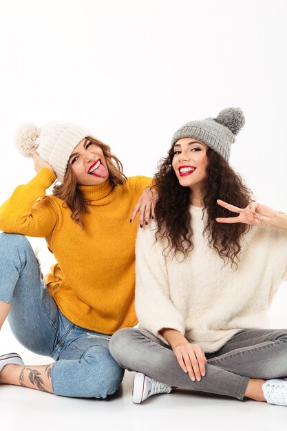 Vertical Two funny girls in sweaters and hats sitting on the floor together while having fun and looking at the camra over white wall