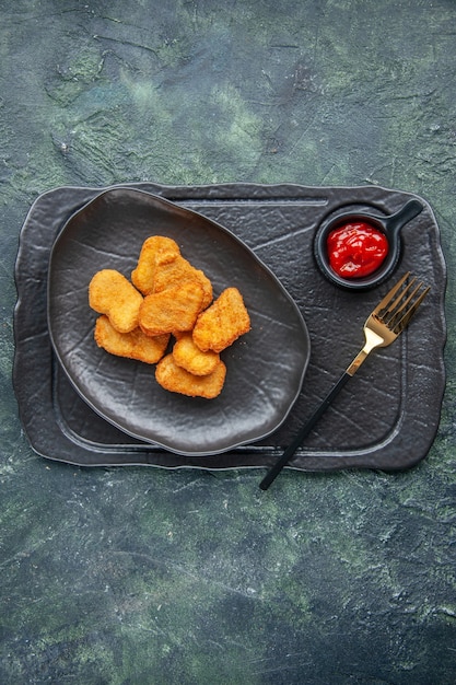 Vertical view of chicken nuggets on a black plate and fork ketchup on dark color tray on dark surface