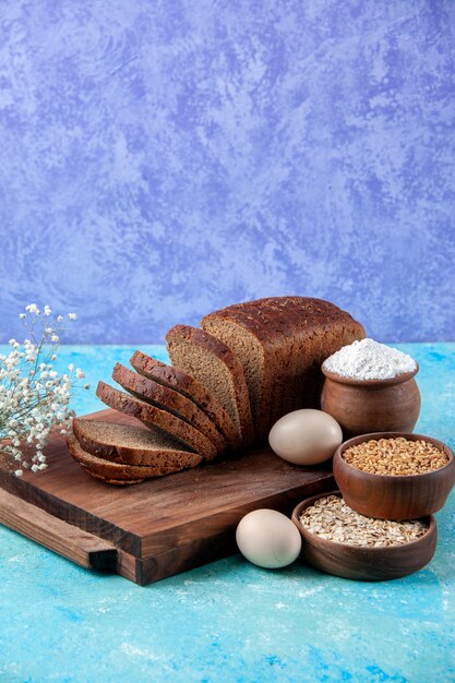 Vertical view of chopped in half black bread slices on wooden boards flour wheat oatmeal in bowls flower eggs on light ice blue pattern background