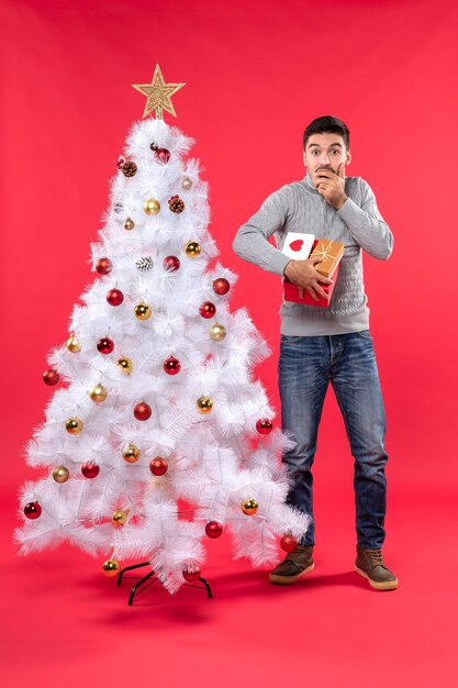 Vertical view of confused handsome man standing near the decorated white xmas tree and holding his gifts on red