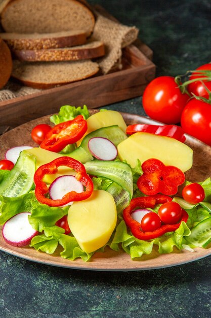 Vertical view of cut fresh black bread on towel in a brown wooden box and chopped fresh vegetables on a plate on dark mix colors surface