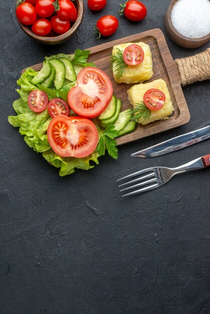 Vertical view of cut fresh tomatoes and cucumbers cheese on wooden board cutlery set salt on black surface