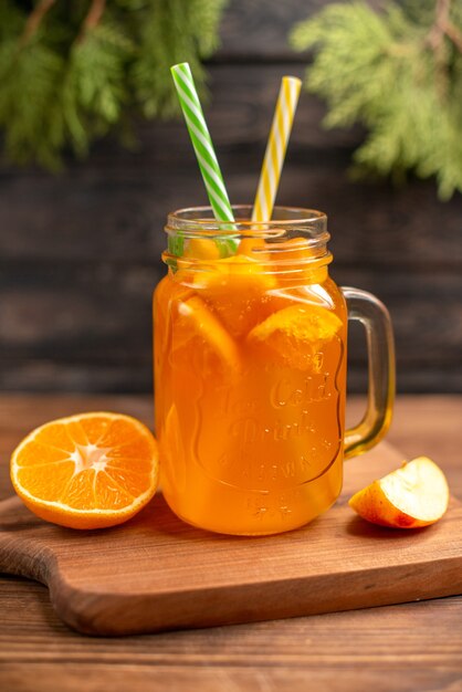 Vertical view of fresh fruit juice in a glass served with tubes and apple and orange on a wooden cutting board on a brown table