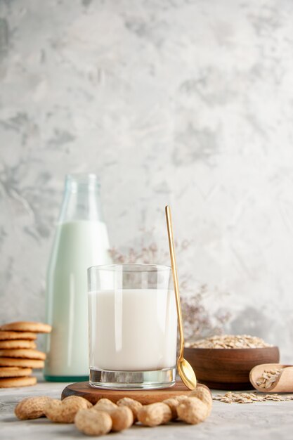 Vertical view of glass bottle and cup filled with milk on wooden tray and dry fruits stacked cookies spoon oats in brown pot on the left side on white table on ice background