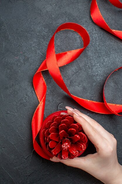 Vertical view of hand holding a conifer cone with red ribbon on dark background