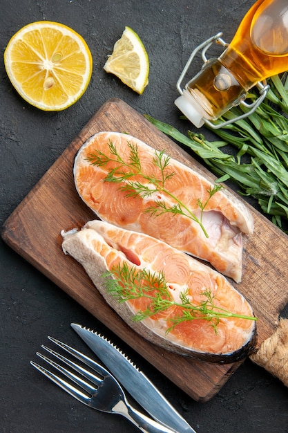 Vertical view of salmon fish on brown wooden cutting board with green lemon slices fallen oil bottle on the right side on dark table